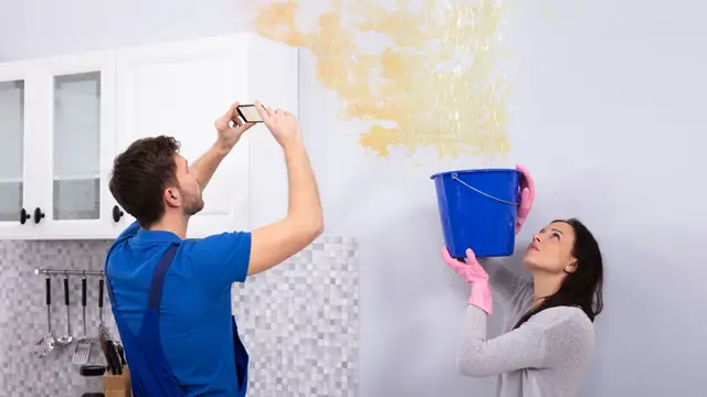 Homeowners photographing a water-stained ceiling while a bucket catches drips