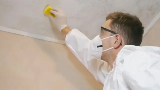 Person in a mask and white suit wiping mold from a ceiling corner